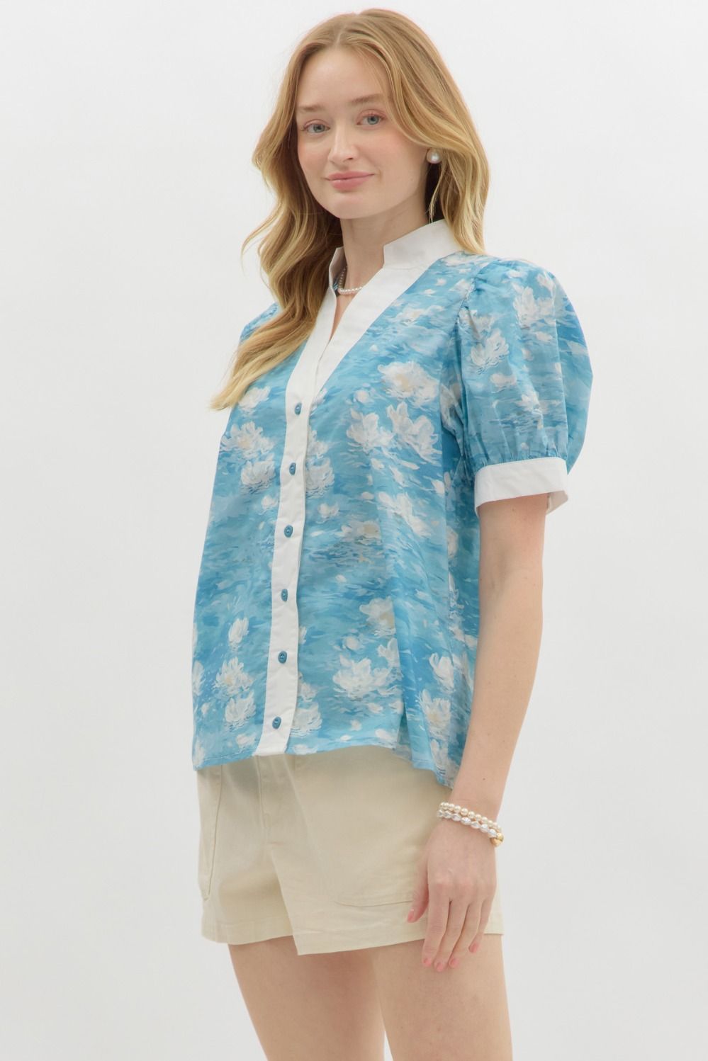 A woman with wavy blonde hair wears a blue floral watercolor puff sleeve blouse with a white mandarin collar and button-front detail. The blouse features a soft, airy fabric with a Monet-inspired water lily print. She pairs it with beige high-waisted shorts and pearl accessories, posing against a clean white background.