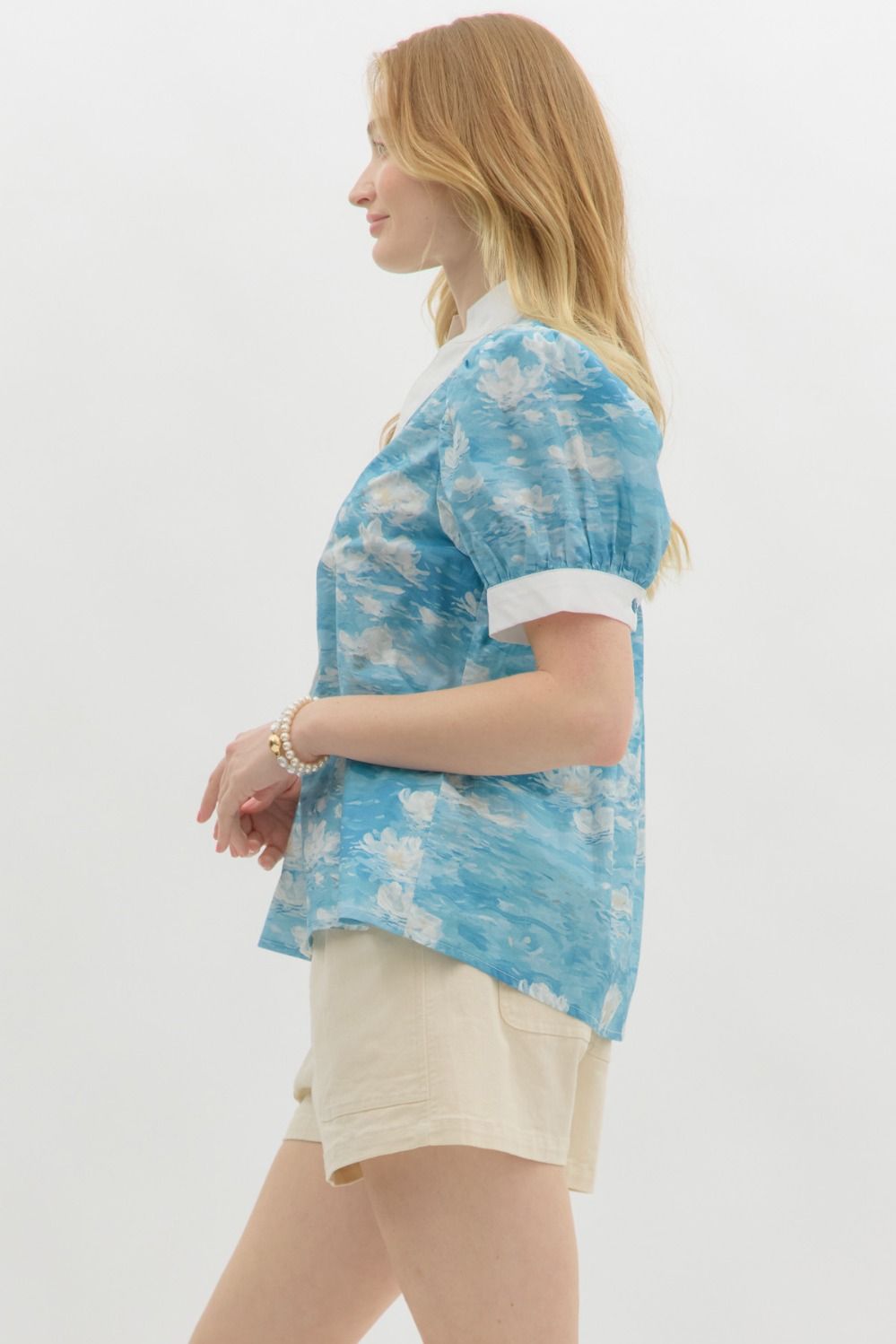 A woman with wavy blonde hair wears a blue floral watercolor puff sleeve blouse with a white mandarin collar and button-front detail. The blouse features a soft, airy fabric with a Monet-inspired water lily print. She pairs it with beige high-waisted shorts and pearl accessories, posing against a clean white background.