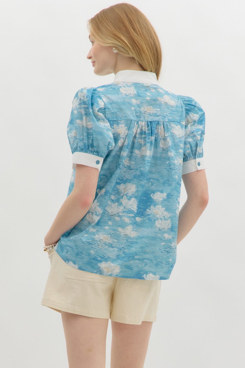 A woman with wavy blonde hair wears a blue floral watercolor puff sleeve blouse with a white mandarin collar and button-front detail. The blouse features a soft, airy fabric with a Monet-inspired water lily print. She pairs it with beige high-waisted shorts and pearl accessories, posing against a clean white background.