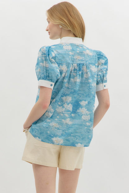 A woman with wavy blonde hair wears a blue floral watercolor puff sleeve blouse with a white mandarin collar and button-front detail. The blouse features a soft, airy fabric with a Monet-inspired water lily print. She pairs it with beige high-waisted shorts and pearl accessories, posing against a clean white background.