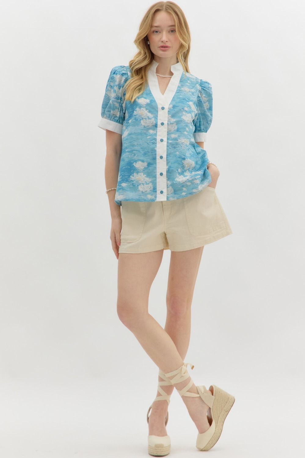 A woman with wavy blonde hair wears a blue floral watercolor puff sleeve blouse with a white mandarin collar and button-front detail. The blouse features a soft, airy fabric with a Monet-inspired water lily print. She pairs it with beige high-waisted shorts and pearl accessories, posing against a clean white background.