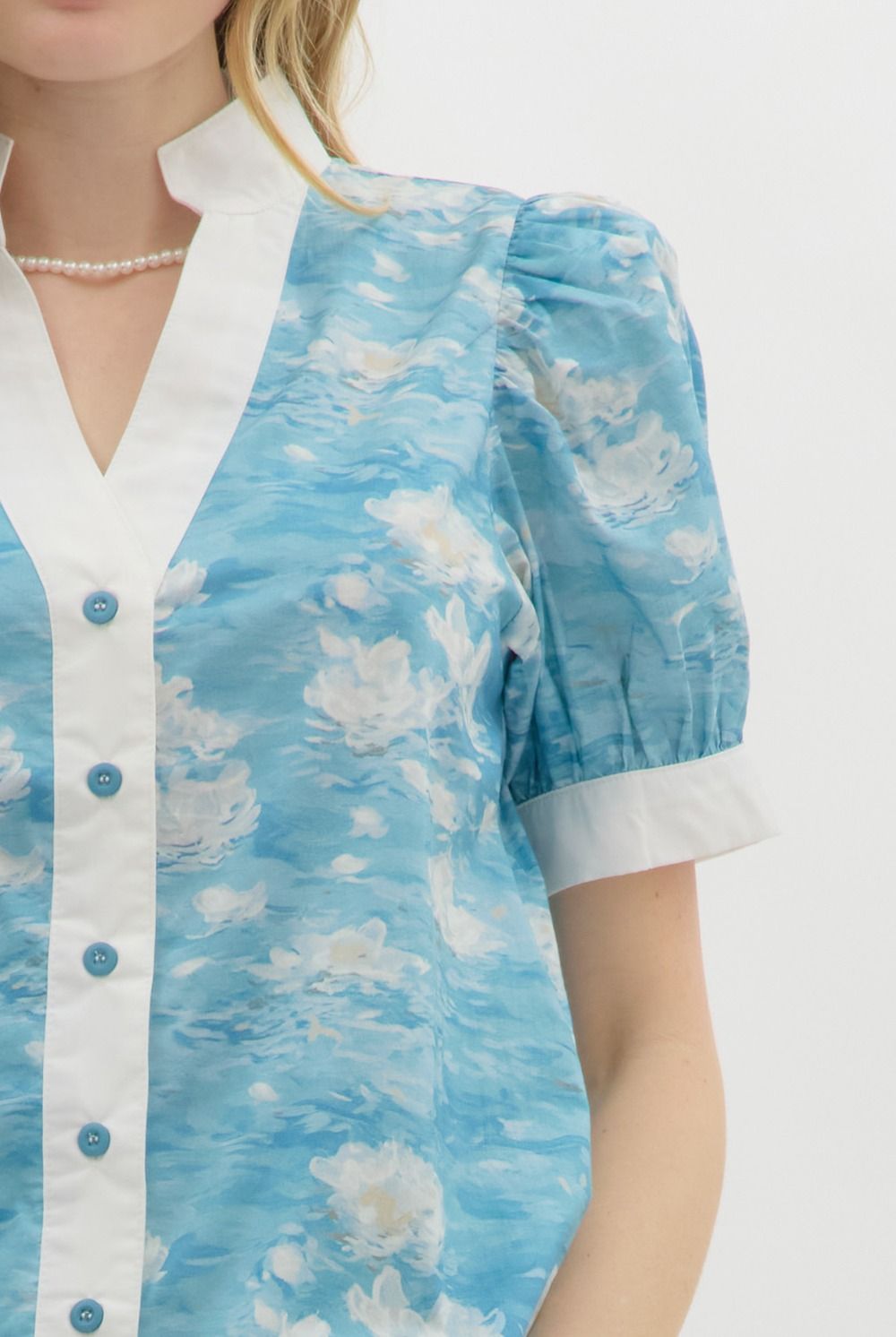 A woman with wavy blonde hair wears a blue floral watercolor puff sleeve blouse with a white mandarin collar and button-front detail. The blouse features a soft, airy fabric with a Monet-inspired water lily print. She pairs it with beige high-waisted shorts and pearl accessories, posing against a clean white background.