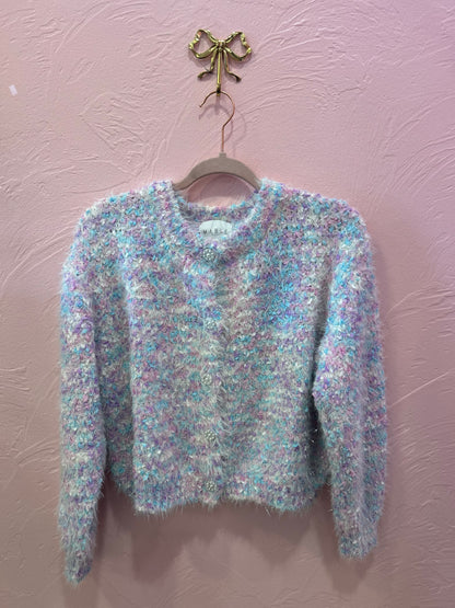 The Frosted Sugar Knit fuzzy pastel cardigan displayed on a hanger against a pink wall, showing soft texture and iridescent button detail.