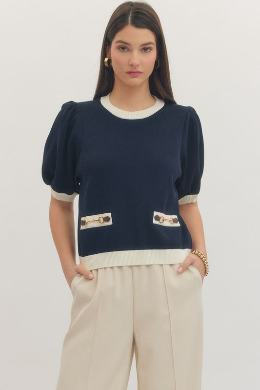 Woman wearing a navy blue top with white trim and beige pants on a plain background
