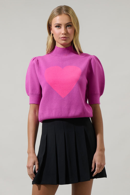Woman wearing a pink sweater with a heart design and black pleated skirt on a gray background