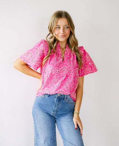 A young woman with wavy blonde hair wearing a hot pink embroidered top with puff sleeves, paired with high-waisted denim jeans. She poses against a neutral background with a soft smile.