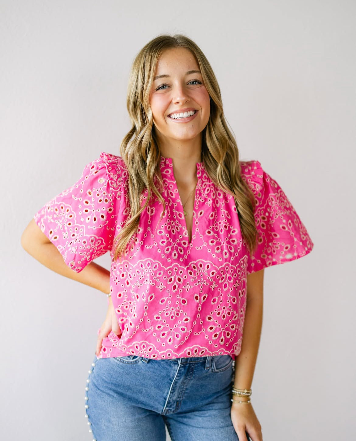 A young woman with wavy blonde hair wearing a hot pink embroidered top with puff sleeves, paired with high-waisted denim jeans. She poses against a neutral background with a soft smile.