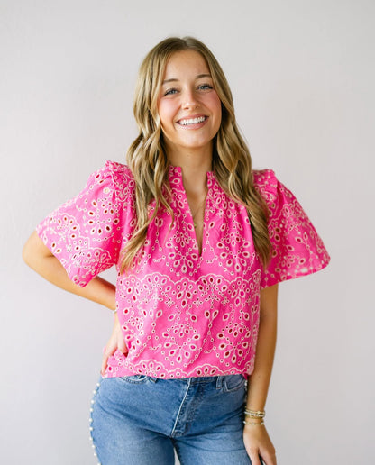 A young woman with wavy blonde hair wearing a hot pink embroidered top with puff sleeves, paired with high-waisted denim jeans. She poses against a neutral background with a soft smile.