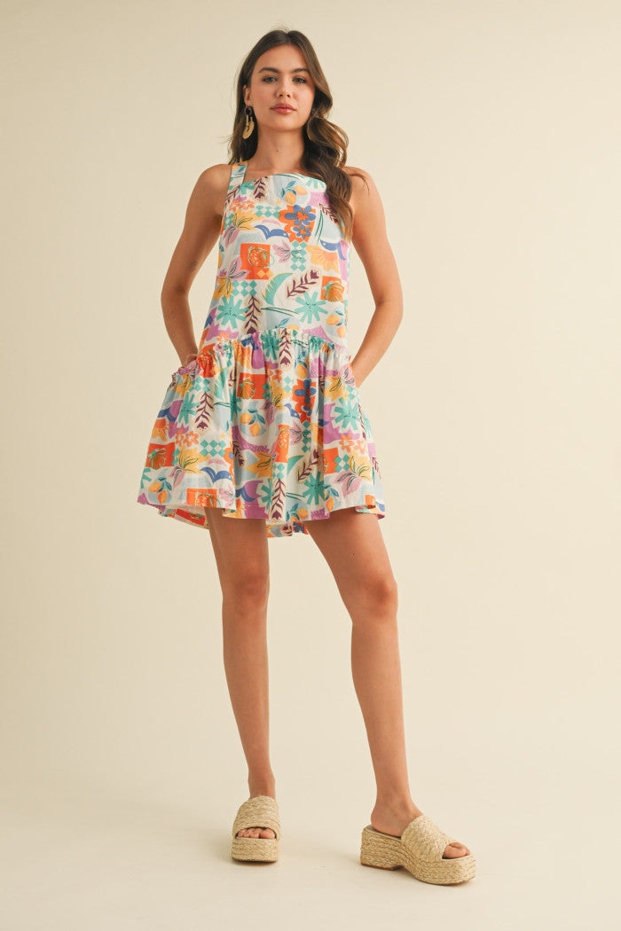 colorful sleeveless sundress with a playful, abstract floral print in shades of orange, purple, turquoise, and pink. The dress features a square neckline and a tiered, flouncy mini skirt. She accessorizes with gold statement earrings and woven platform sandals, standing against a simple beige background