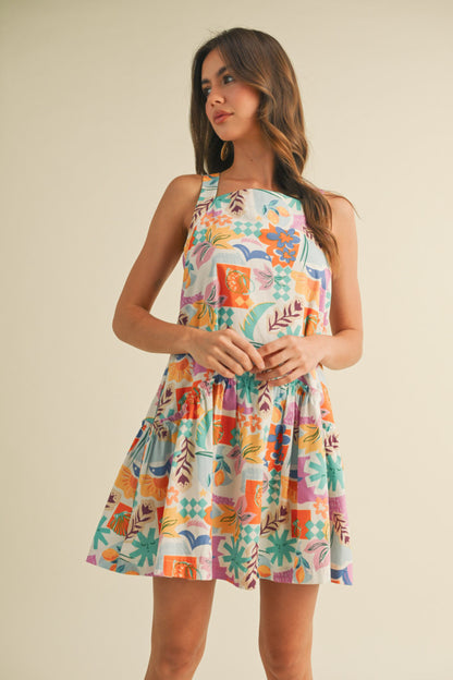 colorful sleeveless sundress with a playful, abstract floral print in shades of orange, purple, turquoise, and pink. The dress features a square neckline and a tiered, flouncy mini skirt. She accessorizes with gold statement earrings and woven platform sandals, standing against a simple beige background