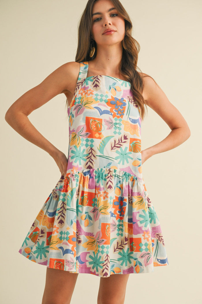 colorful sleeveless sundress with a playful, abstract floral print in shades of orange, purple, turquoise, and pink. The dress features a square neckline and a tiered, flouncy mini skirt. She accessorizes with gold statement earrings and woven platform sandals, standing against a simple beige background