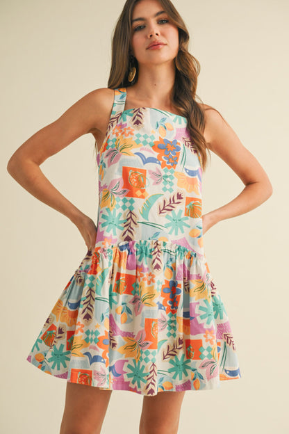colorful sleeveless sundress with a playful, abstract floral print in shades of orange, purple, turquoise, and pink. The dress features a square neckline and a tiered, flouncy mini skirt. She accessorizes with gold statement earrings and woven platform sandals, standing against a simple beige background