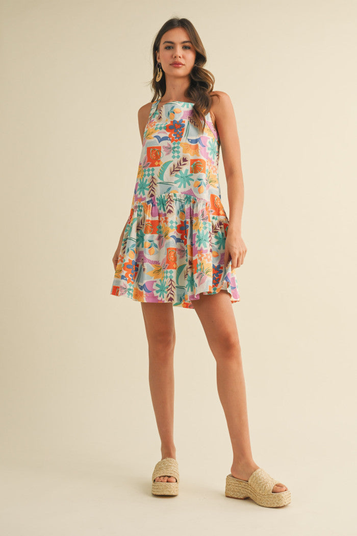 colorful sleeveless sundress with a playful, abstract floral print in shades of orange, purple, turquoise, and pink. The dress features a square neckline and a tiered, flouncy mini skirt. She accessorizes with gold statement earrings and woven platform sandals, standing against a simple beige background