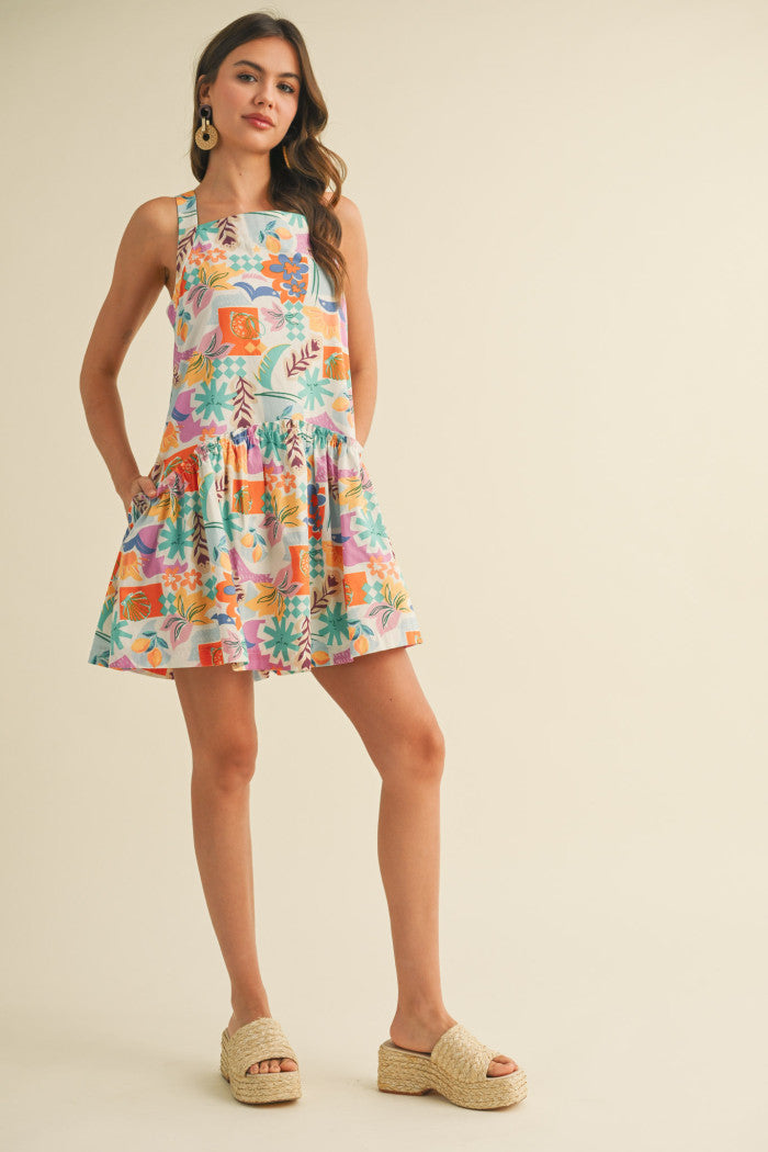 colorful sleeveless sundress with a playful, abstract floral print in shades of orange, purple, turquoise, and pink. The dress features a square neckline and a tiered, flouncy mini skirt. She accessorizes with gold statement earrings and woven platform sandals, standing against a simple beige background