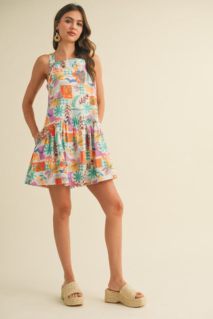 colorful sleeveless sundress with a playful, abstract floral print in shades of orange, purple, turquoise, and pink. The dress features a square neckline and a tiered, flouncy mini skirt. She accessorizes with gold statement earrings and woven platform sandals, standing against a simple beige background