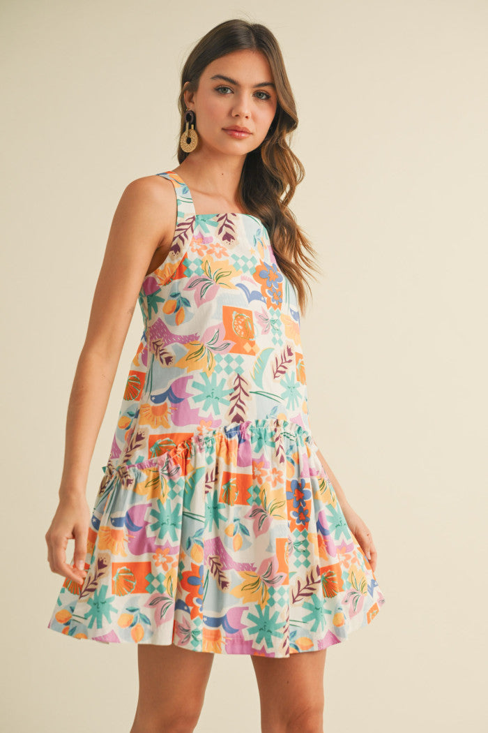colorful sleeveless sundress with a playful, abstract floral print in shades of orange, purple, turquoise, and pink. The dress features a square neckline and a tiered, flouncy mini skirt. She accessorizes with gold statement earrings and woven platform sandals, standing against a simple beige background