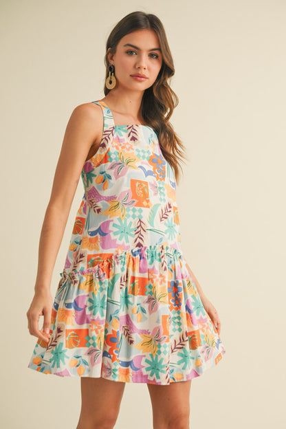 colorful sleeveless sundress with a playful, abstract floral print in shades of orange, purple, turquoise, and pink. The dress features a square neckline and a tiered, flouncy mini skirt. She accessorizes with gold statement earrings and woven platform sandals, standing against a simple beige background