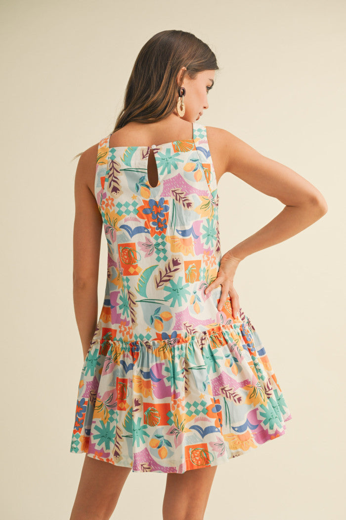 colorful sleeveless sundress with a playful, abstract floral print in shades of orange, purple, turquoise, and pink. The dress features a square neckline and a tiered, flouncy mini skirt. She accessorizes with gold statement earrings and woven platform sandals, standing against a simple beige background