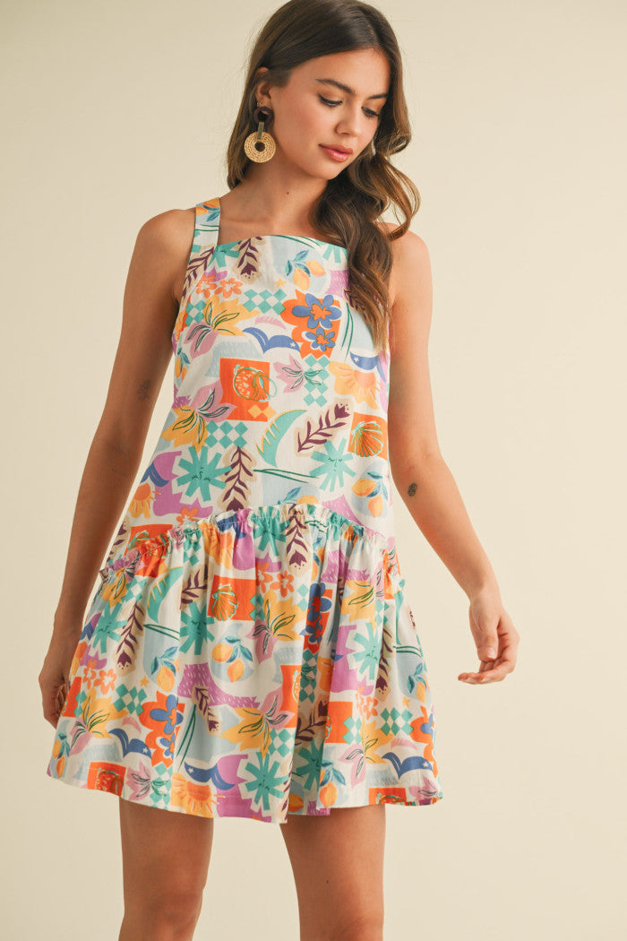 colorful sleeveless sundress with a playful, abstract floral print in shades of orange, purple, turquoise, and pink. The dress features a square neckline and a tiered, flouncy mini skirt. She accessorizes with gold statement earrings and woven platform sandals, standing against a simple beige background