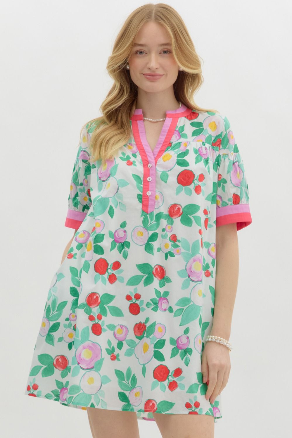 A woman with long blonde hair wears a playful, floral-print mini dress with a relaxed fit. The dress features a white base with vibrant red, pink, and purple flowers, along with green leaves. It has a short-sleeve design with pink contrast trim on the cuffs and neckline, a buttoned V-neck, and a tiered shoulder detail. She accessorizes with pearl jewelry and has a soft, natural smile.