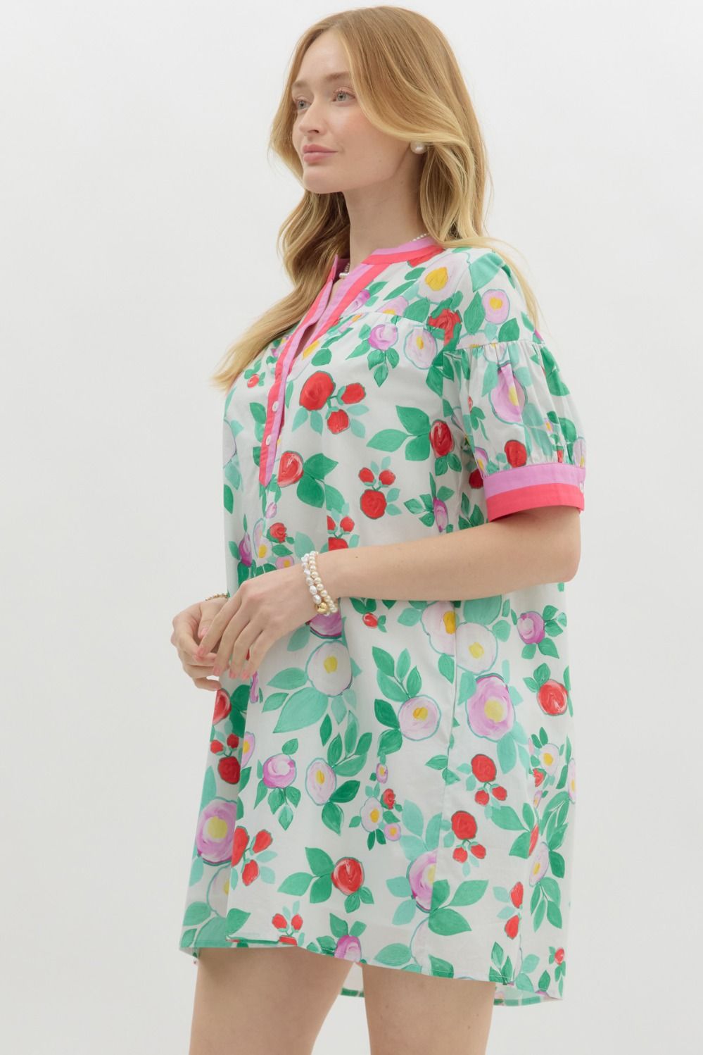 A woman with long blonde hair wears a playful, floral-print mini dress with a relaxed fit. The dress features a white base with vibrant red, pink, and purple flowers, along with green leaves. It has a short-sleeve design with pink contrast trim on the cuffs and neckline, a buttoned V-neck, and a tiered shoulder detail. She accessorizes with pearl jewelry and has a soft, natural smile.