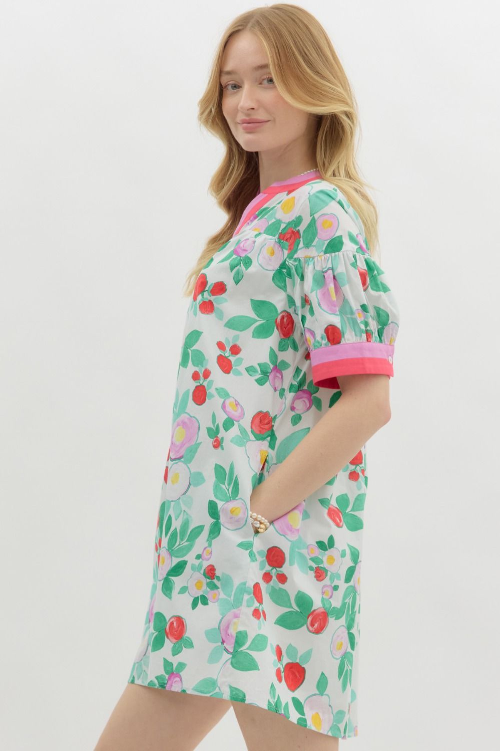 A woman with long blonde hair wears a playful, floral-print mini dress with a relaxed fit. The dress features a white base with vibrant red, pink, and purple flowers, along with green leaves. It has a short-sleeve design with pink contrast trim on the cuffs and neckline, a buttoned V-neck, and a tiered shoulder detail. She accessorizes with pearl jewelry and has a soft, natural smile.