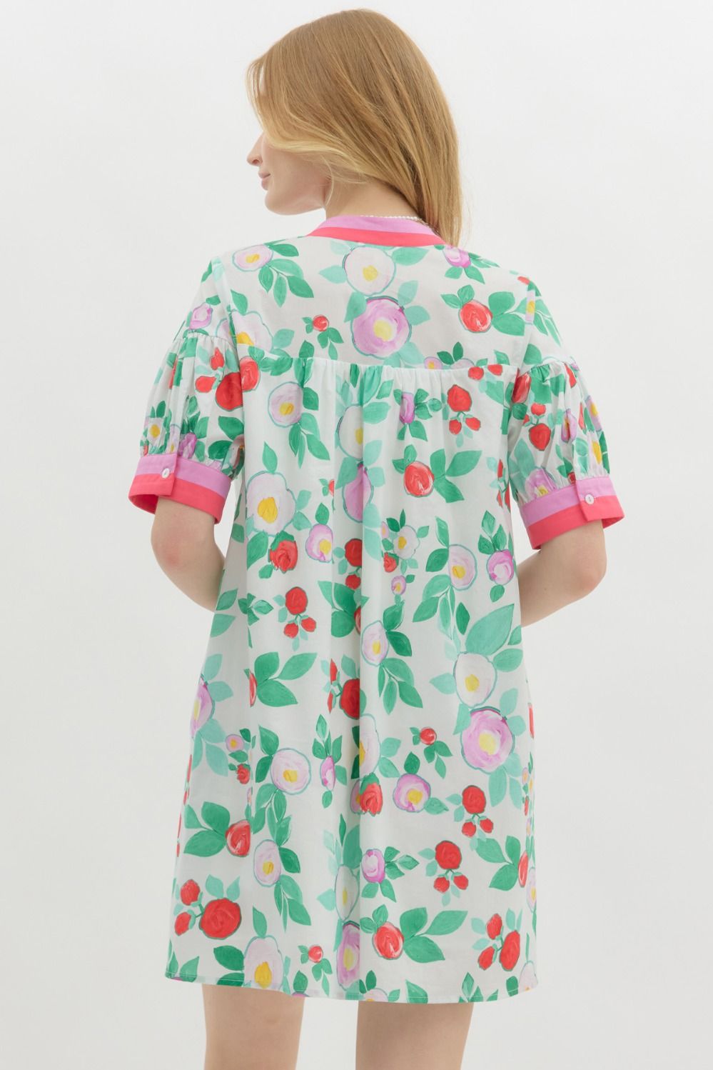 A woman with long blonde hair wears a playful, floral-print mini dress with a relaxed fit. The dress features a white base with vibrant red, pink, and purple flowers, along with green leaves. It has a short-sleeve design with pink contrast trim on the cuffs and neckline, a buttoned V-neck, and a tiered shoulder detail. She accessorizes with pearl jewelry and has a soft, natural smile.