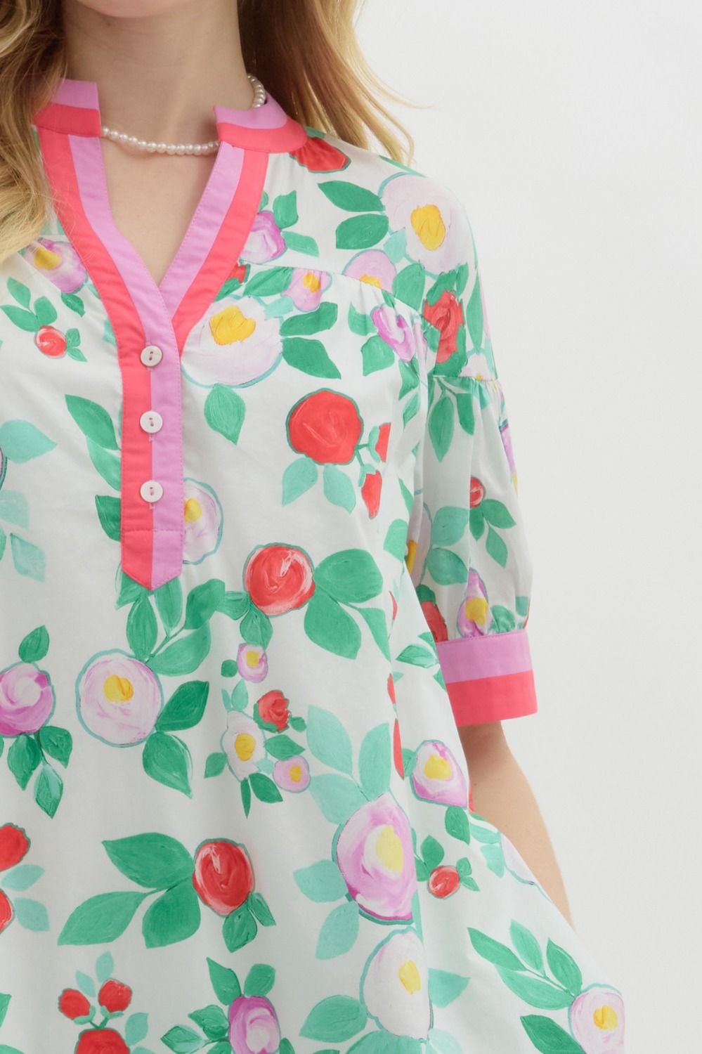 A woman with long blonde hair wears a playful, floral-print mini dress with a relaxed fit. The dress features a white base with vibrant red, pink, and purple flowers, along with green leaves. It has a short-sleeve design with pink contrast trim on the cuffs and neckline, a buttoned V-neck, and a tiered shoulder detail. She accessorizes with pearl jewelry and has a soft, natural smile.