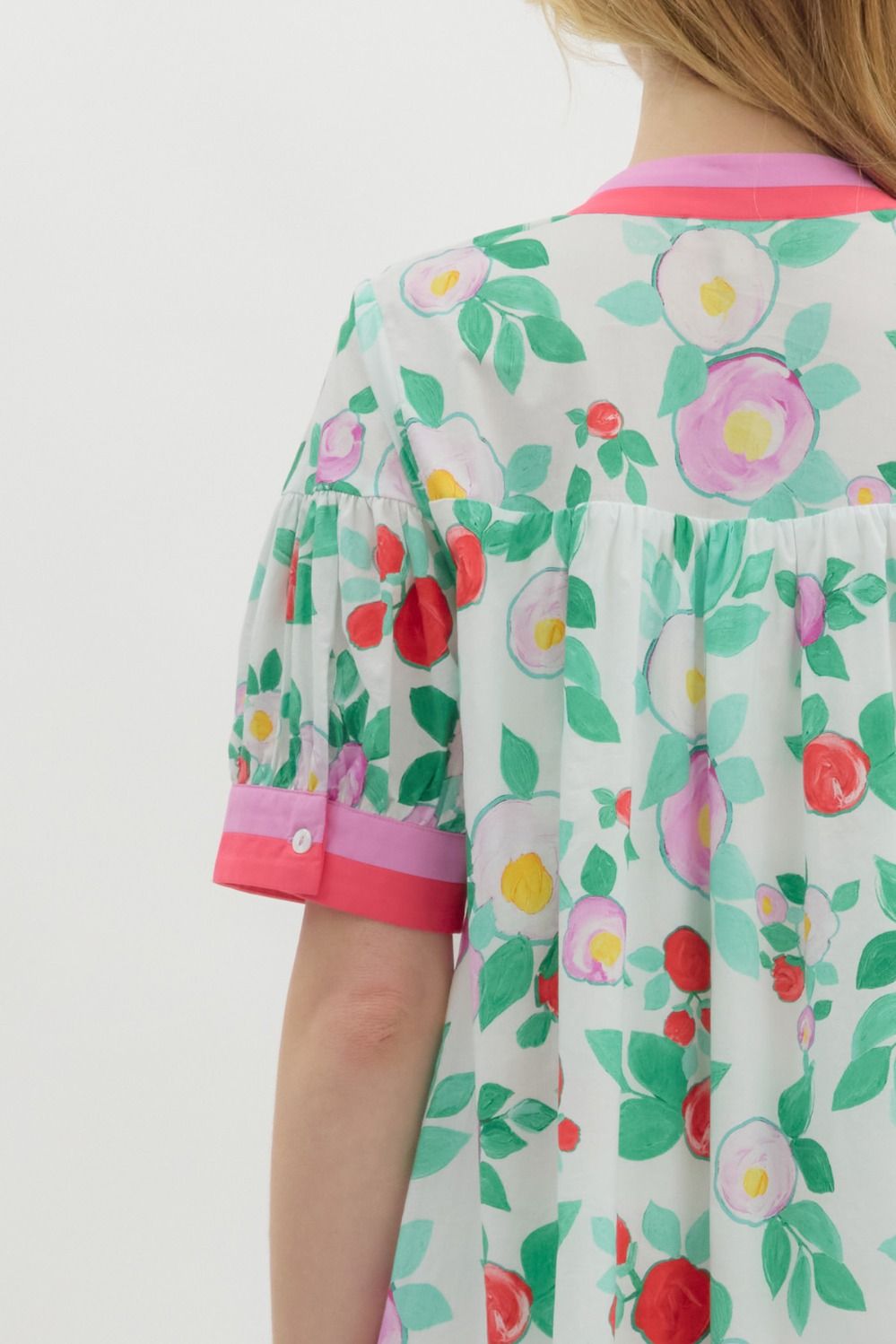 A woman with long blonde hair wears a playful, floral-print mini dress with a relaxed fit. The dress features a white base with vibrant red, pink, and purple flowers, along with green leaves. It has a short-sleeve design with pink contrast trim on the cuffs and neckline, a buttoned V-neck, and a tiered shoulder detail. She accessorizes with pearl jewelry and has a soft, natural smile.