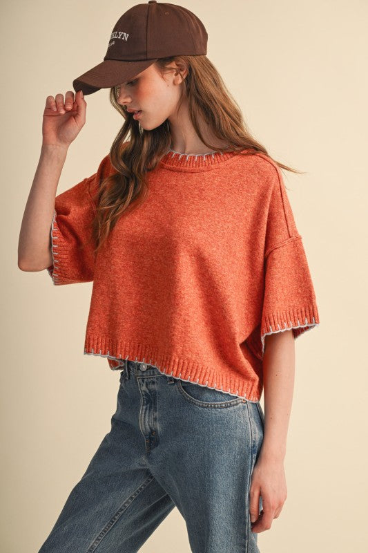 Front view of rust orange cropped short sleeve sweater with pale blue contrast stitch edging.