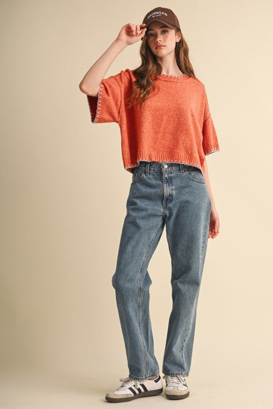 Full-length outfit with orange and blue contrast stitch sweater, blue jeans and sneakers.