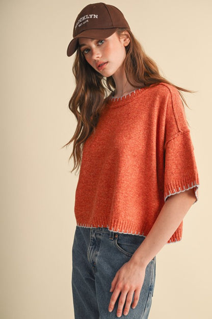 Rust orange short-sleeve knit sweater with light blue contrast stitching at neckline, cuffs and hem, side view.