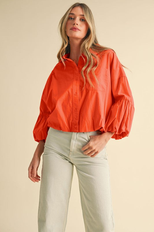 Model wearing bright orange button-front blouse with voluminous balloon sleeves and collared neckline.