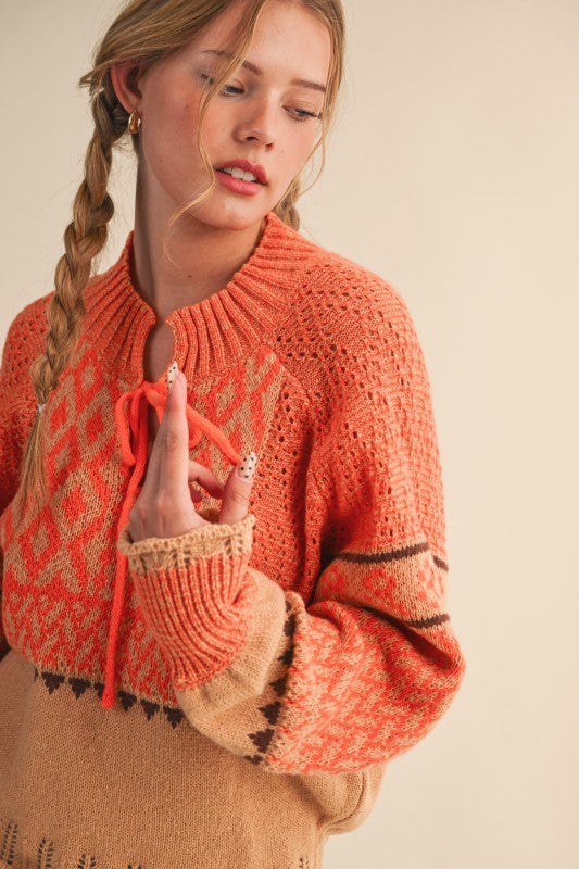 Close view of orange tie-neck sweater with textured knit shoulders and patterned sleeves.