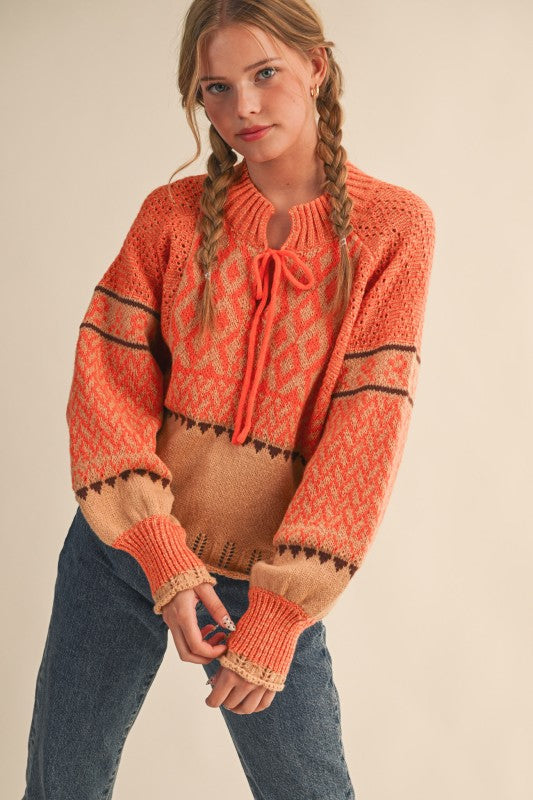 Front view of orange and tan patterned knit sweater with tie neckline and long sleeves.