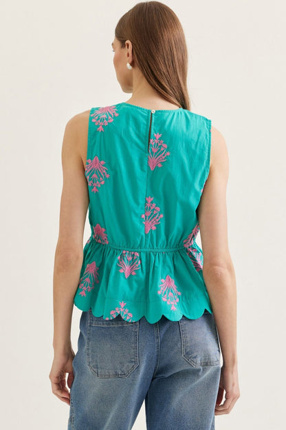 Woman wearing a teal top with pink floral embroidery and blue jeans on a beige background