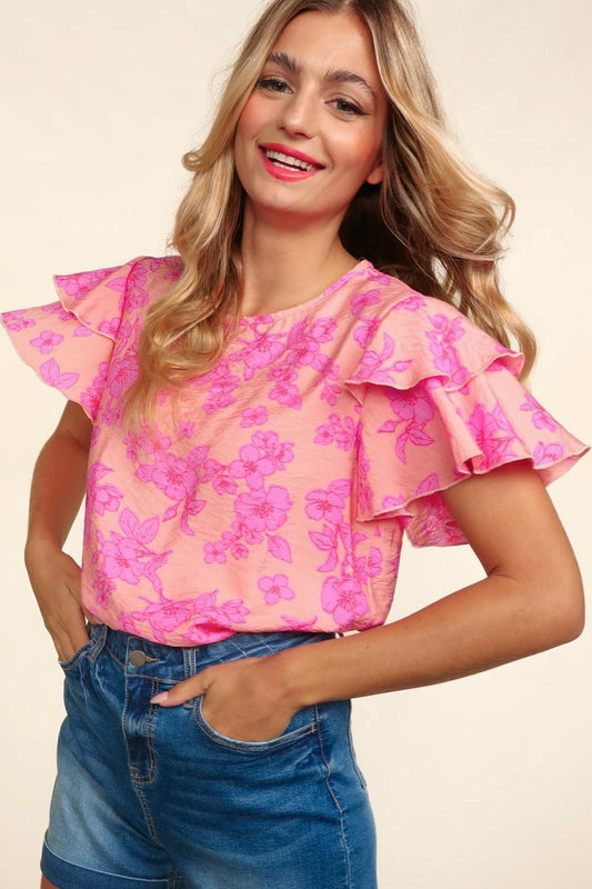 Woman wearing a peach-pink floral top with double ruffle sleeves and merrow edge detailing. Styled with high-waisted denim shorts for a playful and feminine look.