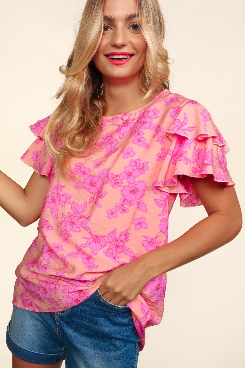 Woman wearing a peach-pink floral top with double ruffle sleeves and merrow edge detailing. Styled with high-waisted denim shorts for a playful and feminine look.
