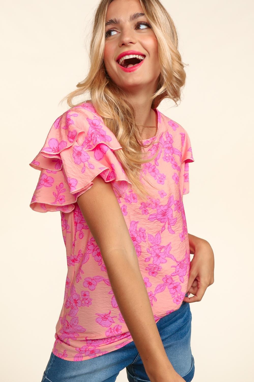 Woman wearing a peach-pink floral top with double ruffle sleeves and merrow edge detailing. Styled with high-waisted denim shorts for a playful and feminine look.