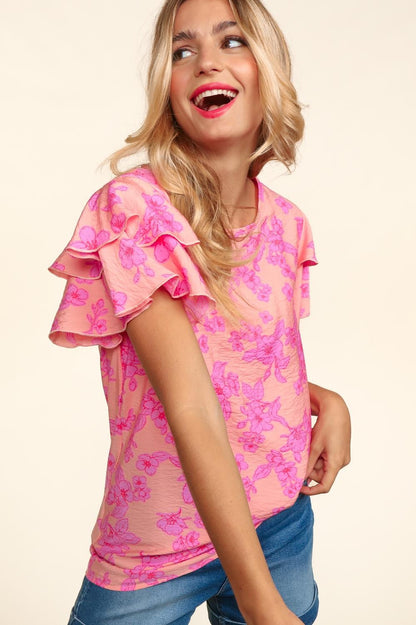 Woman wearing a peach-pink floral top with double ruffle sleeves and merrow edge detailing. Styled with high-waisted denim shorts for a playful and feminine look.