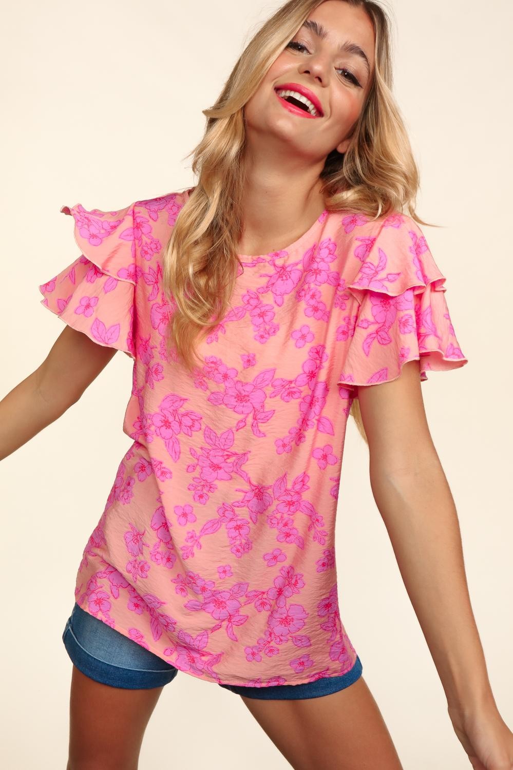 Woman wearing a peach-pink floral top with double ruffle sleeves and merrow edge detailing. Styled with high-waisted denim shorts for a playful and feminine look.