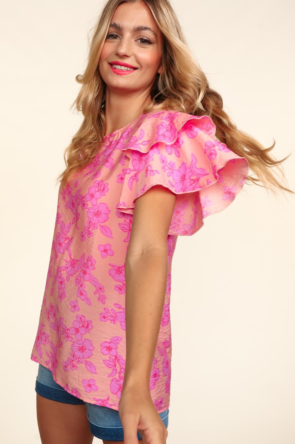 Woman wearing a peach-pink floral top with double ruffle sleeves and merrow edge detailing. Styled with high-waisted denim shorts for a playful and feminine look.