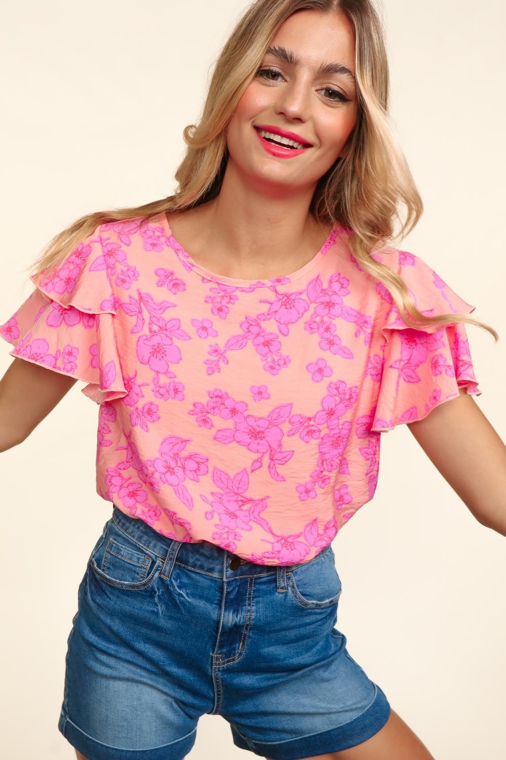 Woman wearing a peach-pink floral top with double ruffle sleeves and merrow edge detailing. Styled with high-waisted denim shorts for a playful and feminine look.