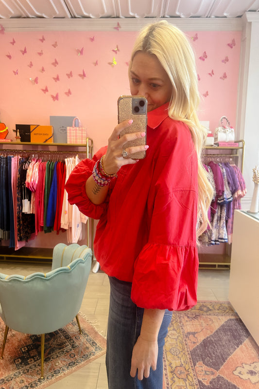 Woman taking a mirror selfie wearing a red blouse in a store with pink walls and butterfly decorations.