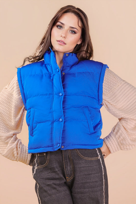 A woman wearing a bright Thunder Blue puffer vest styled over a textured long-sleeve top and dark denim jeans.