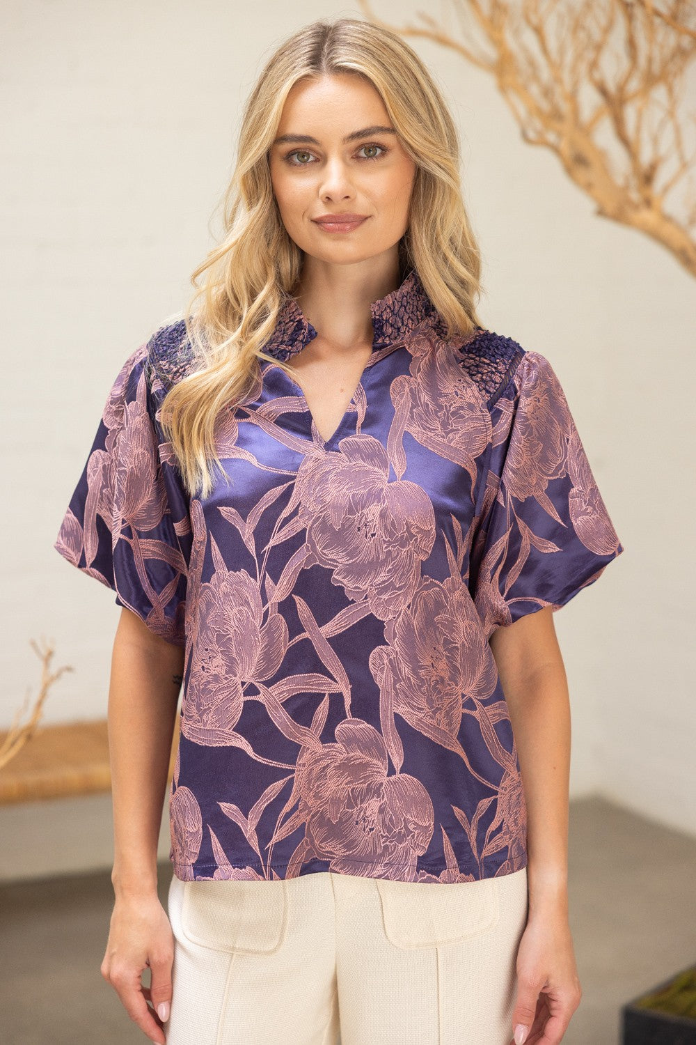 Model wearing The Twilight Bloom Top — a violet satin blouse with blush floral pattern, smocked shoulder detail, and split neckline.