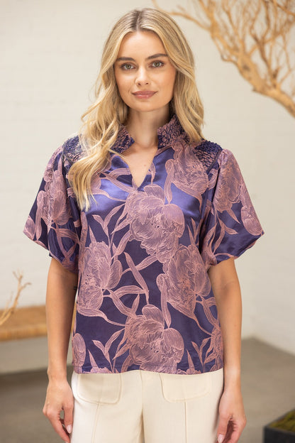 Model wearing The Twilight Bloom Top — a violet satin blouse with blush floral pattern, smocked shoulder detail, and split neckline.