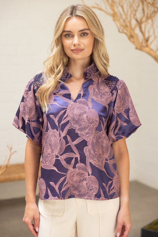 Model wearing The Twilight Bloom Top — a violet satin blouse with blush floral pattern, smocked shoulder detail, and split neckline.